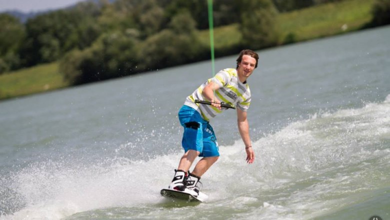 A wakeboarder rides on a lake surrounded by a green landscape.