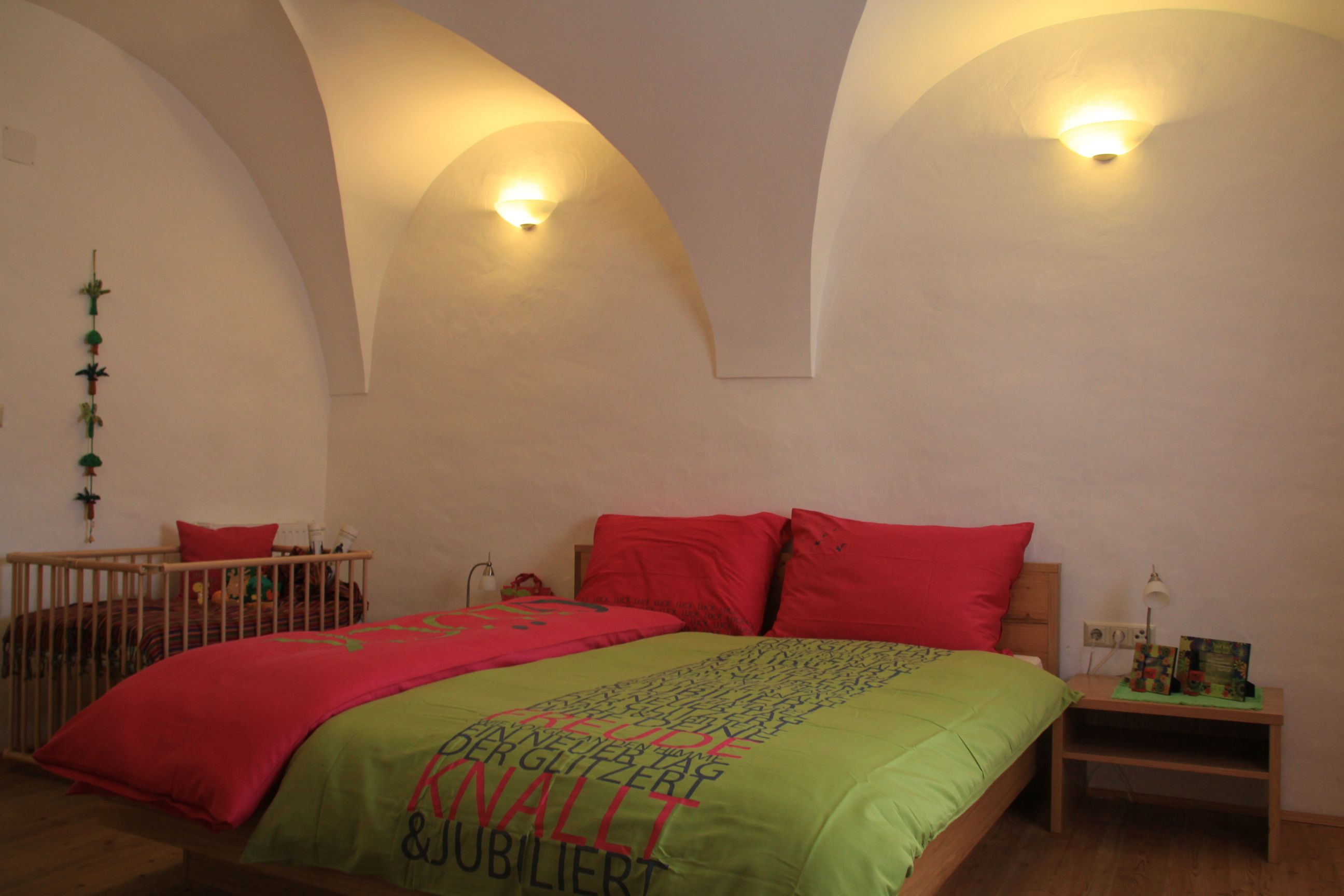 A cozy guest room with a double bed, colorful comforter covers and a baby crib in the background. The walls are white and curved.