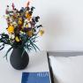 A table with a black vase full of colorful flowers, a blue magazine with the inscription 'HOLLA!' and a white cushion.