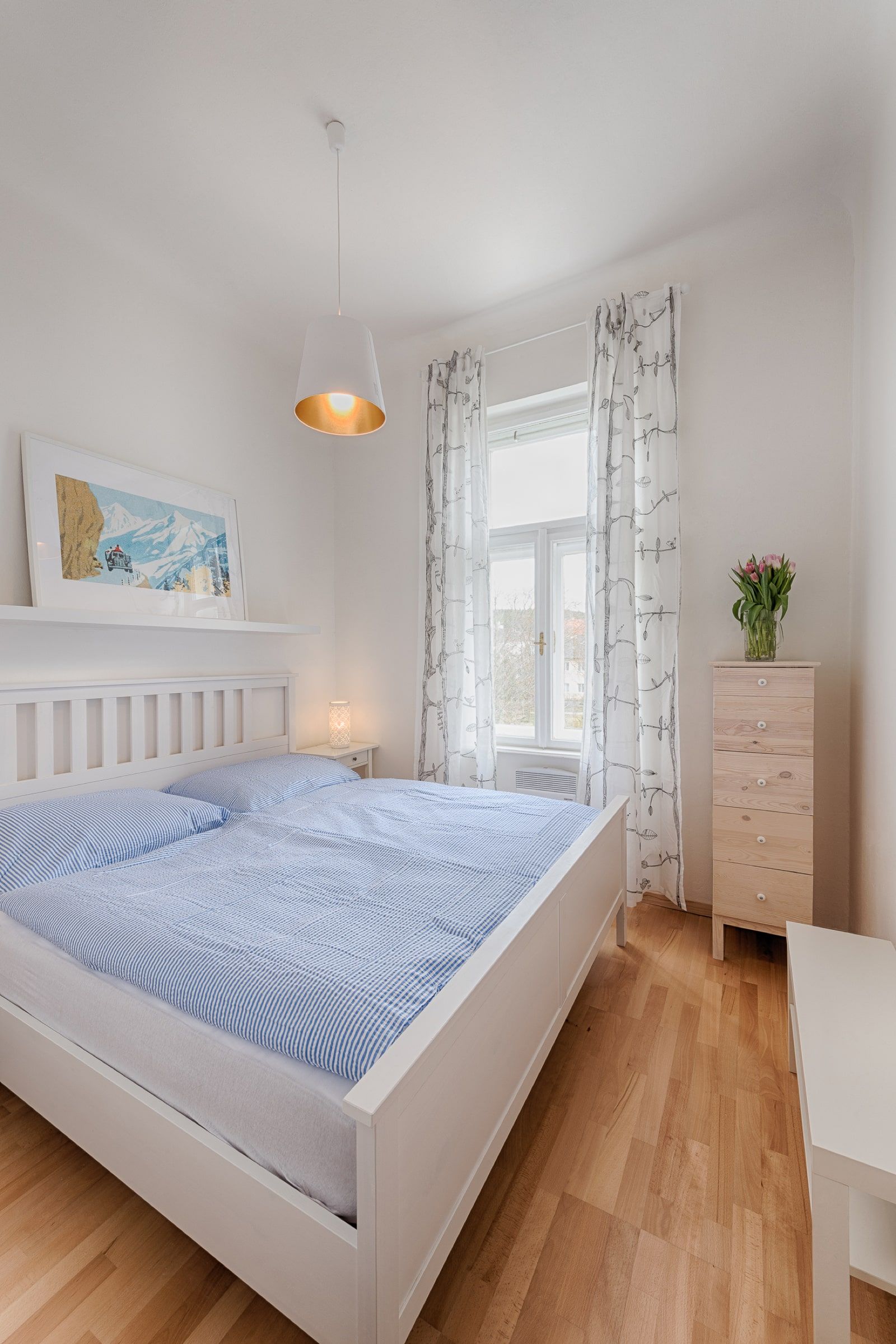 Bright bedroom with white bed, blue bed linen, wooden chest of drawers and window with curtains.