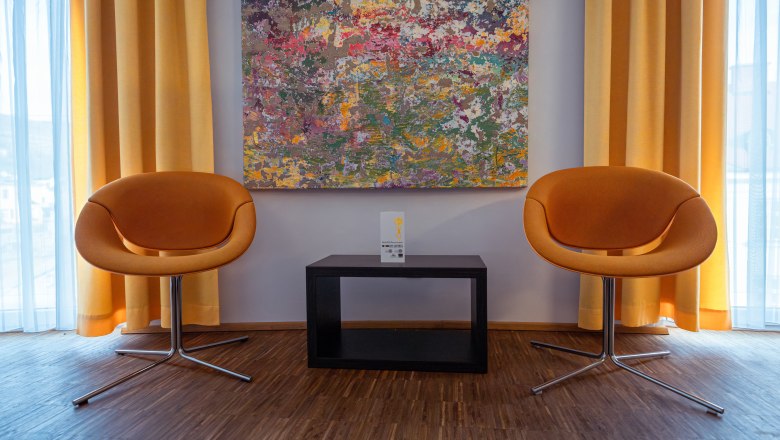 Two orange chairs in front of a colorful painting and yellow curtains.