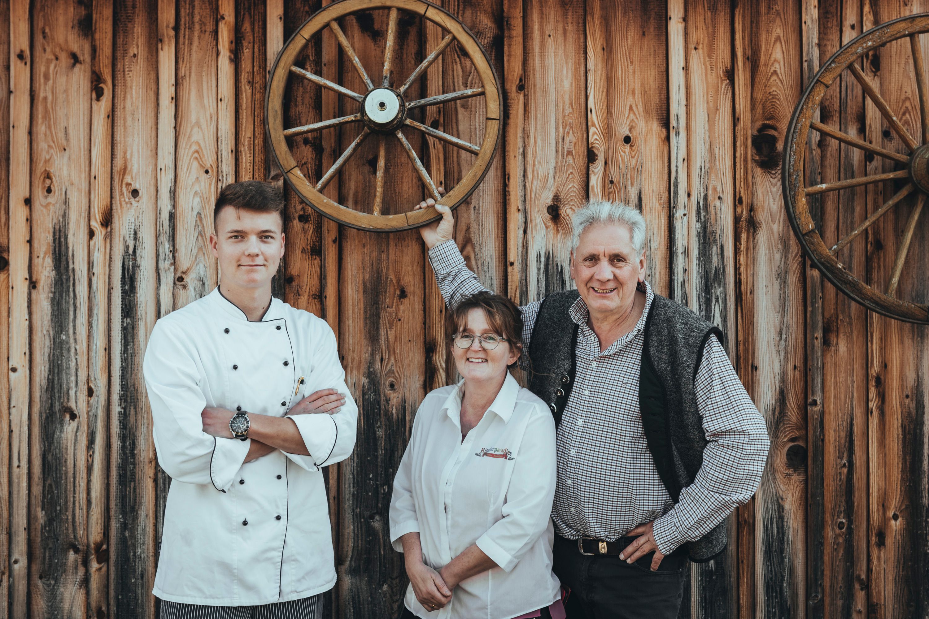 Three people in front of a wooden wall with wagon wheels, one person in cooking clothes.