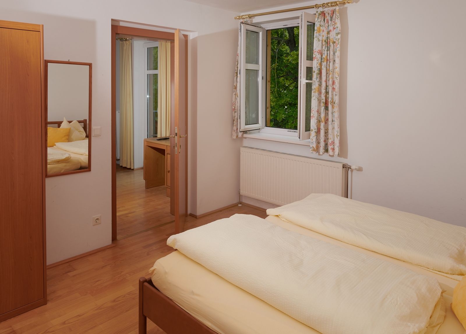 A bedroom with a double bed, an open window with curtains and a mirror on the wall.