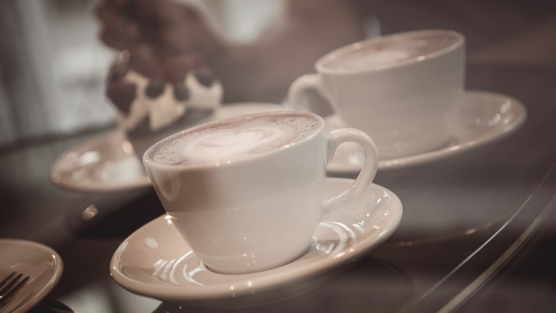 Two cups of coffee on saucers, a dessert in the background.