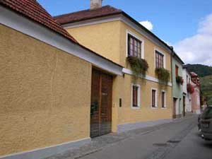 House, © Emmerich Granner Yellow house with red roof tiles and flower boxes on the windows in a narrow street.