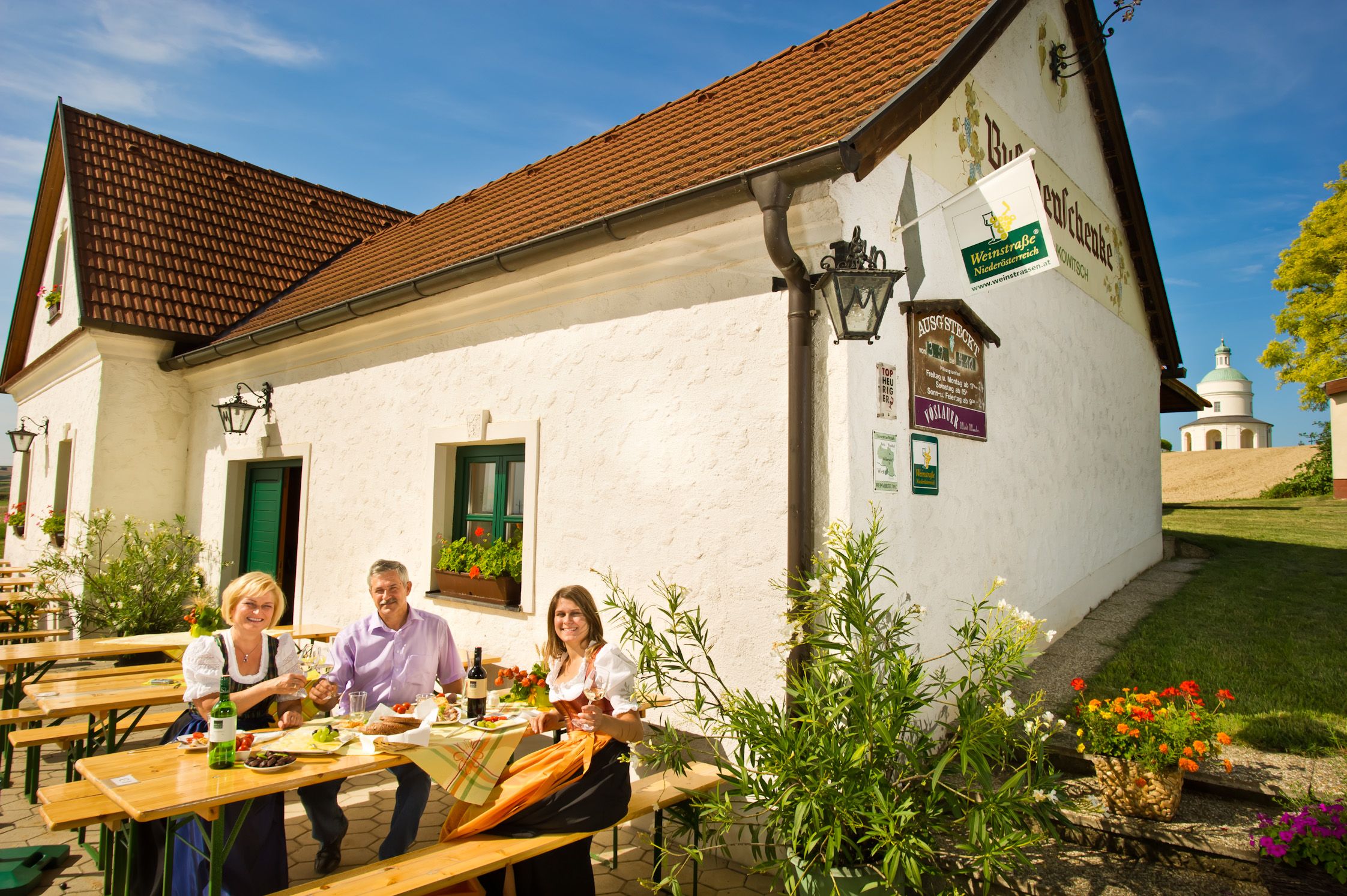 Three people sit at a table in front of a winery and enjoy a meal outdoors.