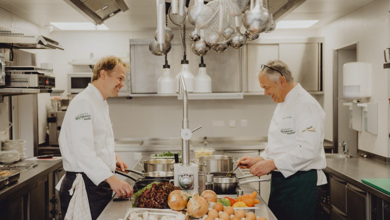 Two chefs in a professional kitchen cooking together. There are vegetables and eggs on the table.