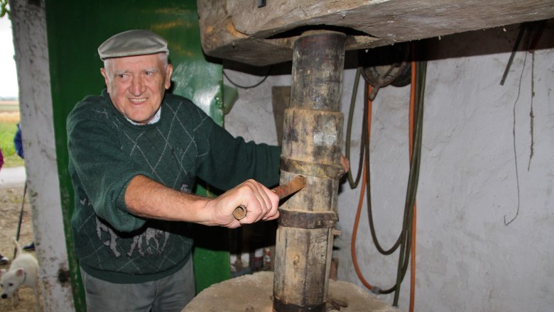 An elderly man in a cap operates an old mechanical press in a rustic room.