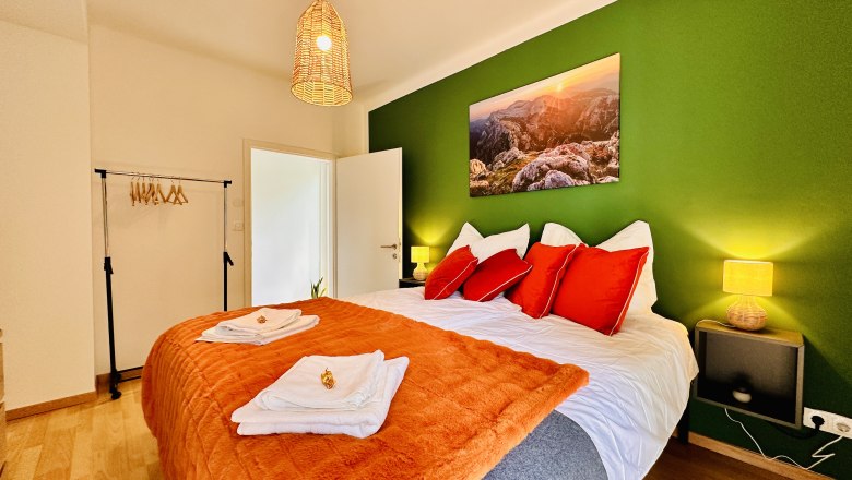 Cozy bedroom with green painted wall, double bed with red pillows, orange blanket and landscape picture. Hanging lamp and clothes rack available.