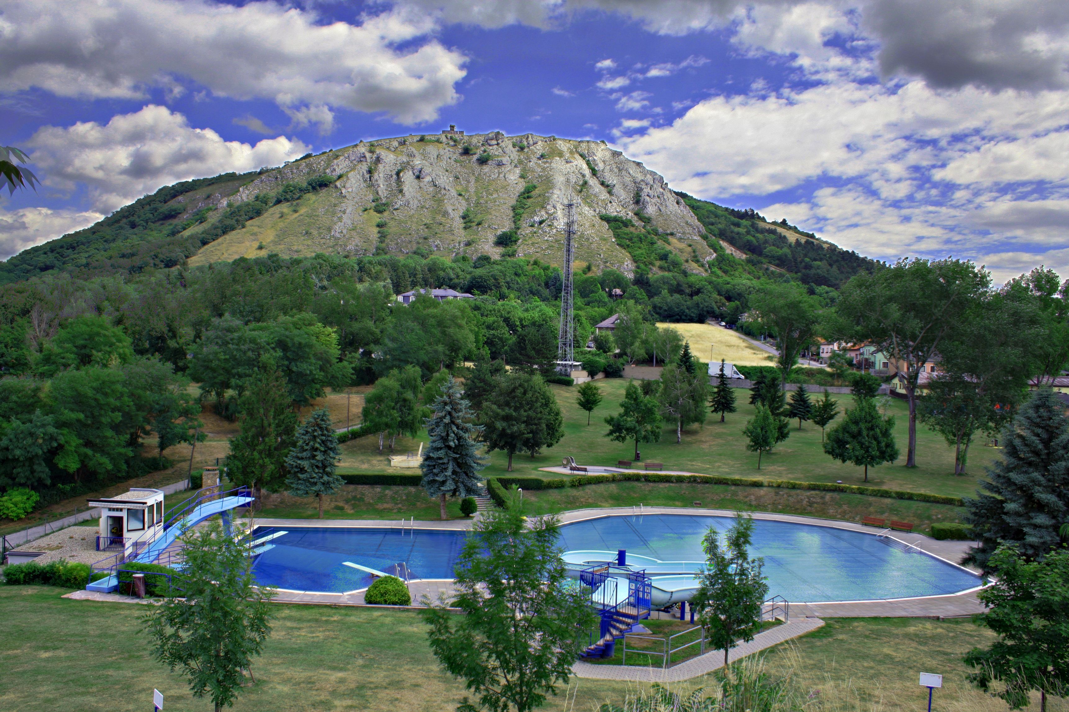 An outdoor pool with a round pool and a slide, surrounded by trees, in front of a wooded hill with a rocky peak.
