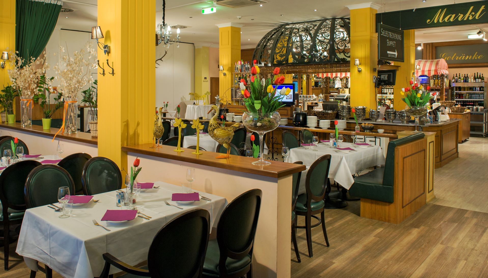 Restaurant with tables separated by half walls and yellow concrete columns. The kitchen is discreetly visible in the background.