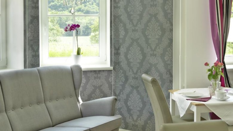 Country house Smöch, © Familie Smoech Interior of a stylish living room with gray sofa, decorative wallpaper, chandelier and dining table with flowers.