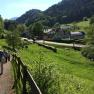 People walk along a path in a green, hilly landscape with houses in the background.