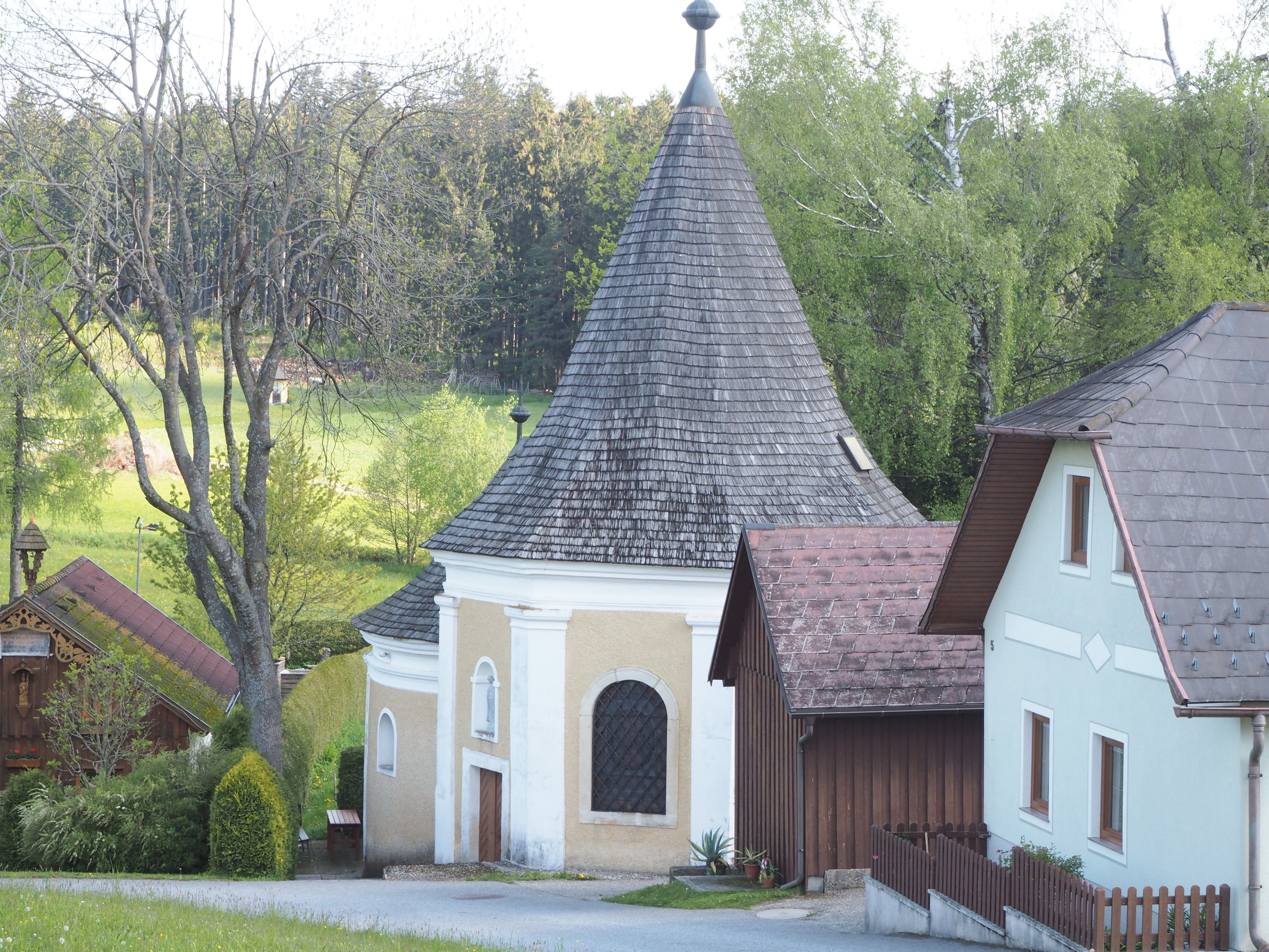 A small chapel with a pointed roof, surrounded by trees and houses.