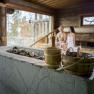 Interior view of a sauna with two people sitting on wooden benches in the background. Sauna buckets and stones can be seen in the foreground.