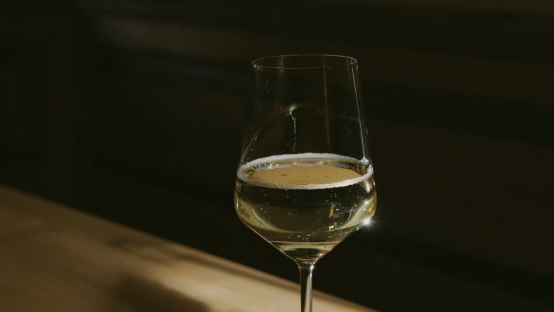 A wine glass with white wine stands on a wooden table in the shade.