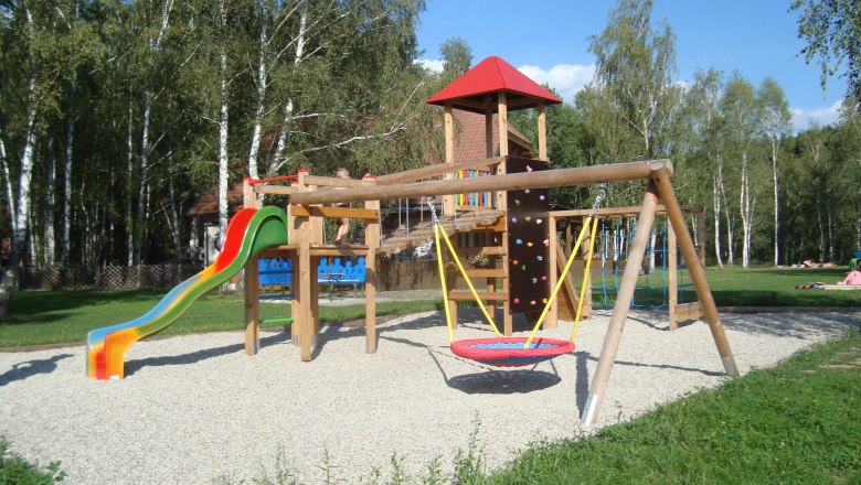 A children's playground with slide, swings and climbing frame, surrounded by trees.