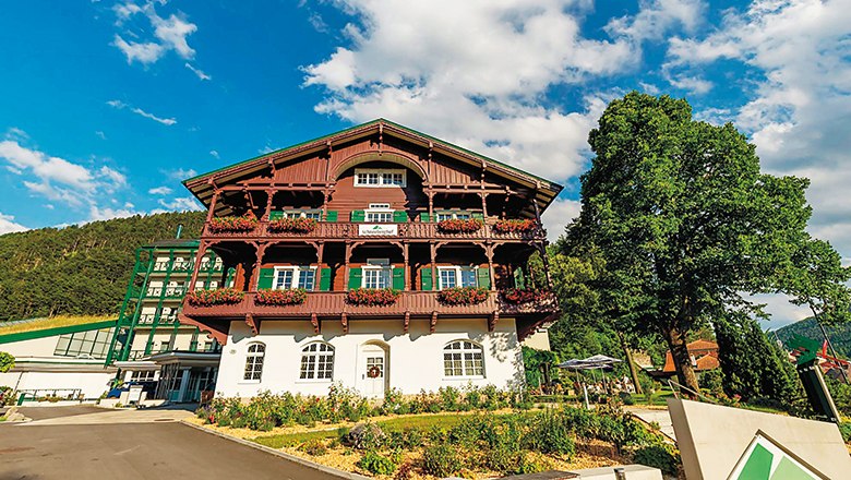 A traditional, multi-storey building with wooden balconies and flowers, surrounded by trees and a blue sky.