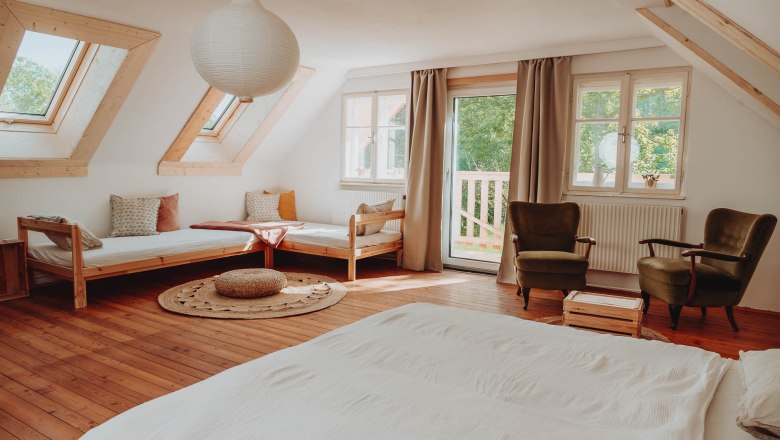 Bright multi-bed room with wooden floor, two beds, two armchairs and large windows.