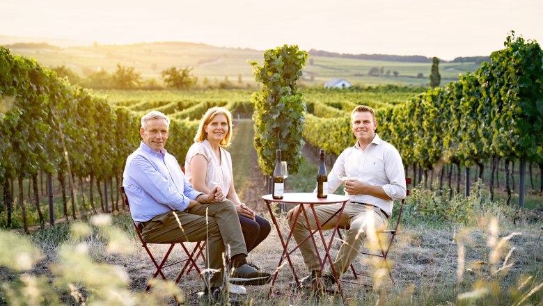 Three people sit at a table in the vineyard with wine glasses and bottles.
