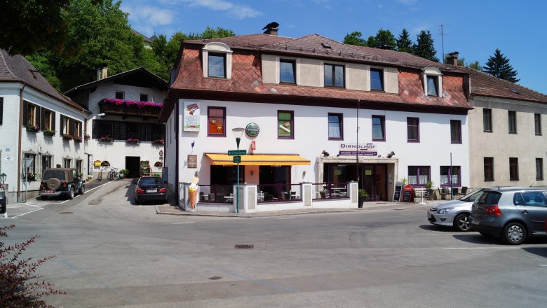 A building with the sign 'Dirndlhof' and a terrace with a parasol. Cars are parked in the parking lot in front of it.