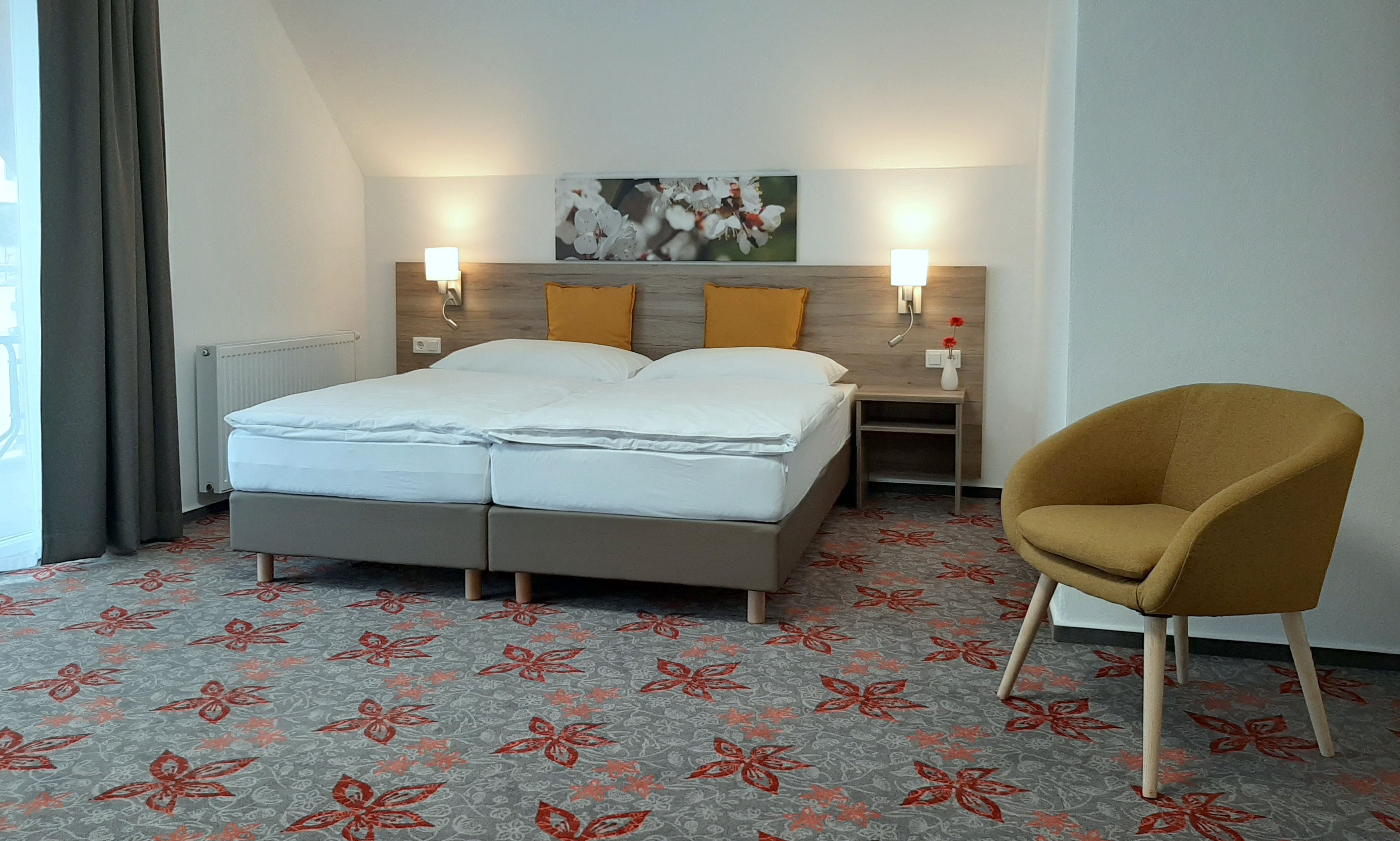 Modern double room with two beds, yellow cushions, carpet with floral pattern and a yellow armchair.