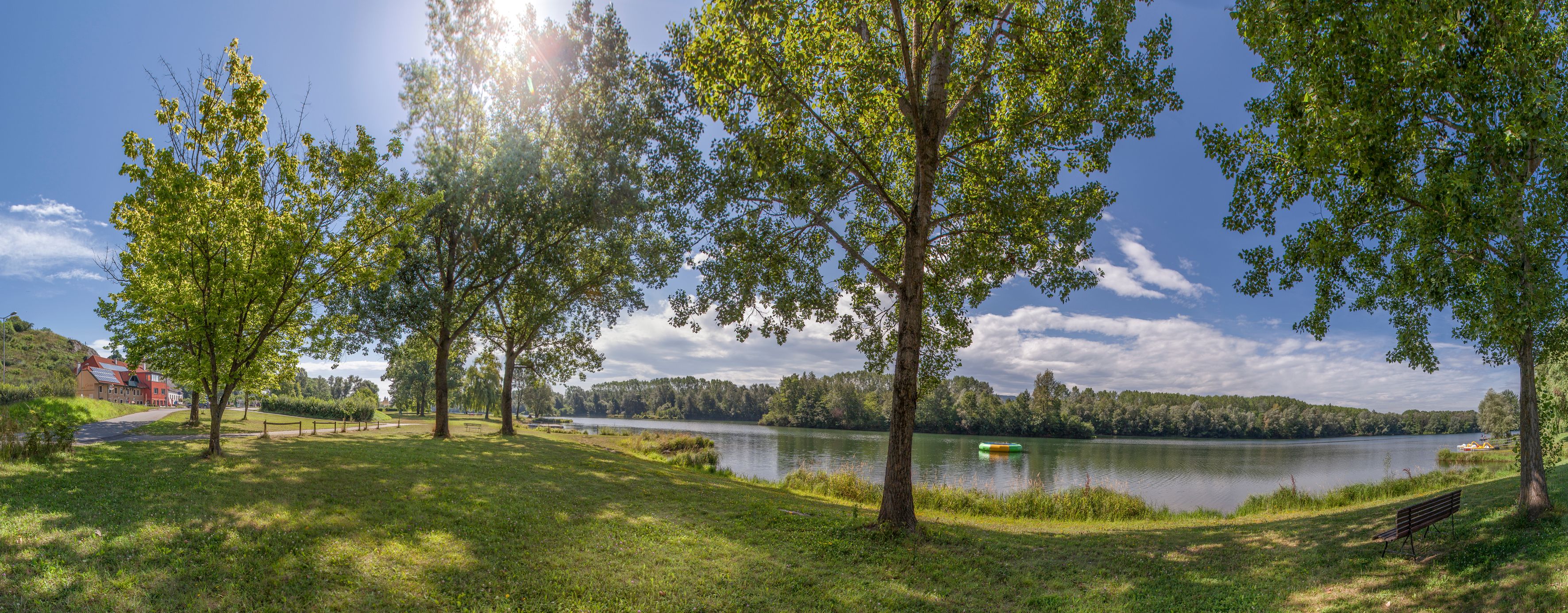 Panoramic view of a lake with trees and a bench in the foreground, sunny sky.