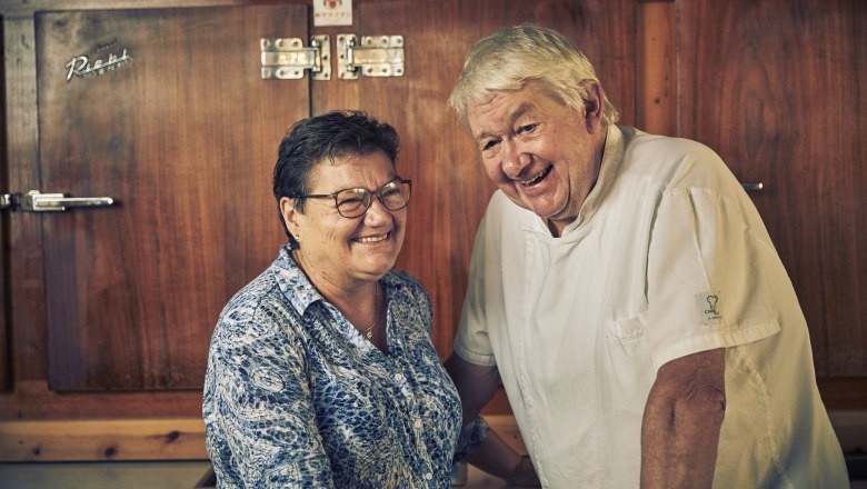 An elderly couple smiles in a kitchen. The man is wearing a chef's jacket, the woman a patterned blouse. They are standing in front of a wooden cupboard.