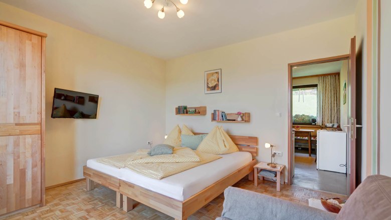 A cozy bedroom with a wooden floor, a double bed with yellow bed linen, a flat-screen TV on the wall and an open passageway to the kitchen.