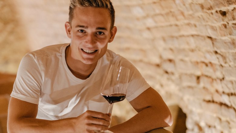 A young man sits on a wine barrel in a cellar and holds a glass of red wine in his hand.