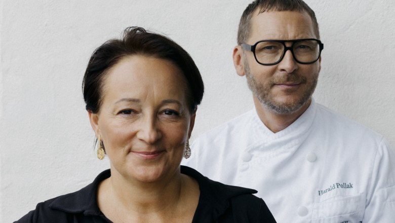 Two people are standing in front of a white wall. A woman in black and a man in chef's clothes.