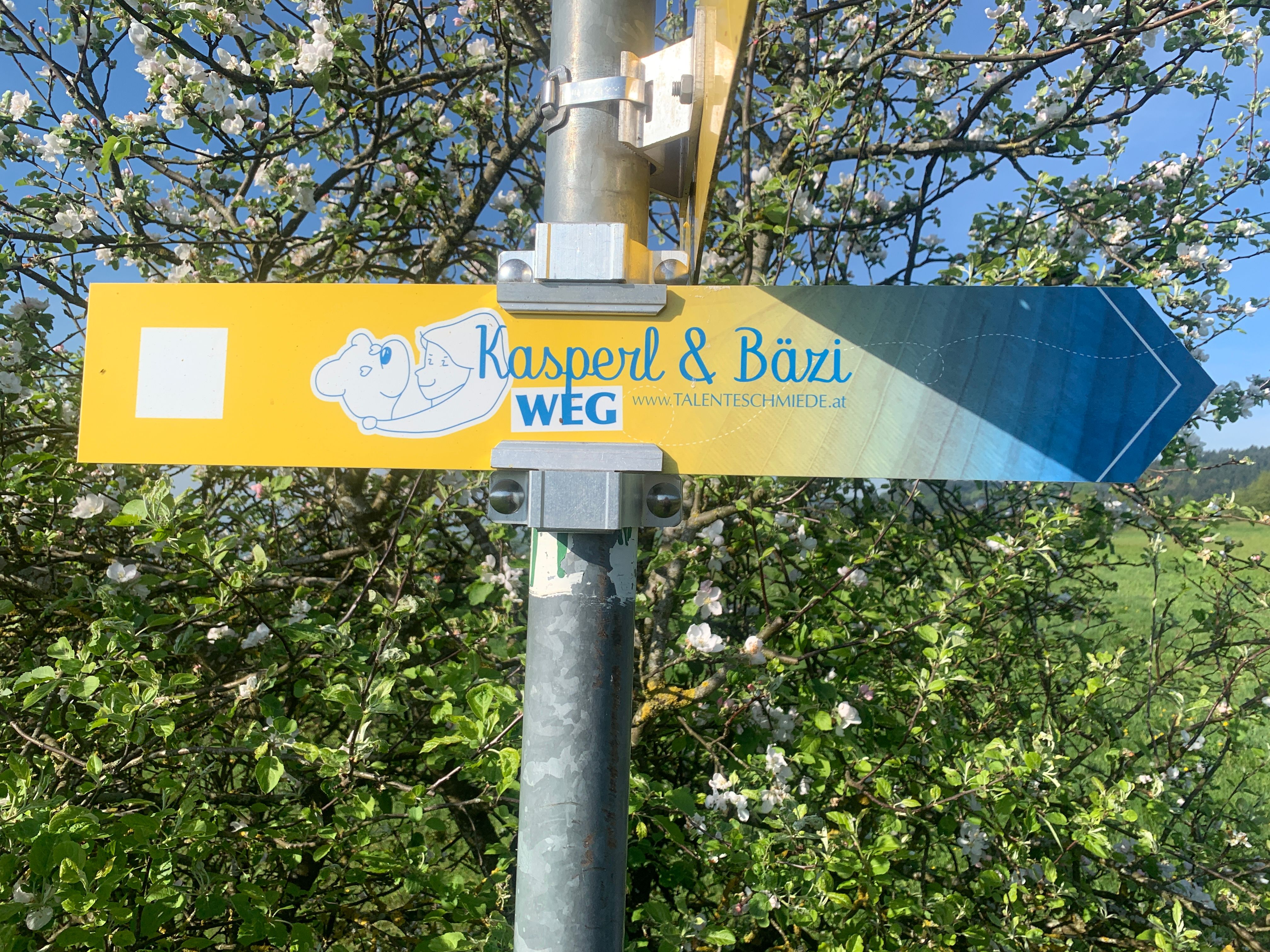 Signpost with the inscription 'Kasperl & Bäzi WEG' in front of a blossoming tree.