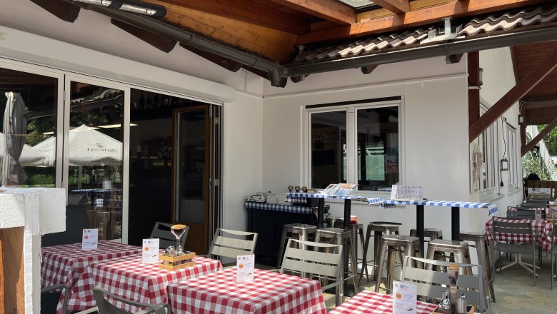 Entrance area of the pizzeria with guest garden. In the foreground are set tables with red tablecloths.