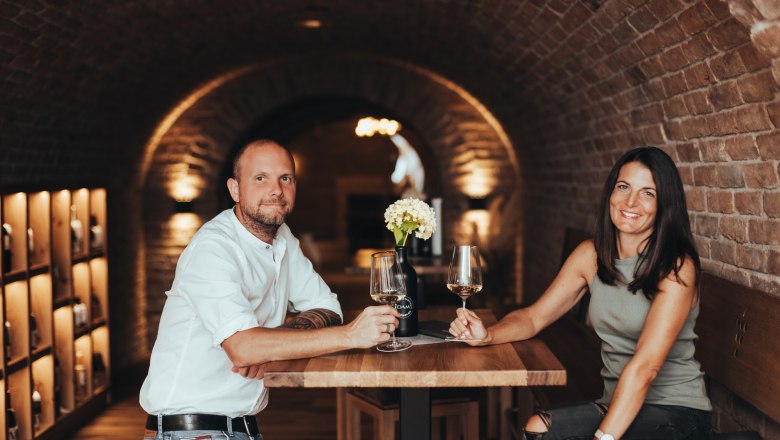 A man and a woman are sitting at a table in a wine cellar holding glasses of wine.