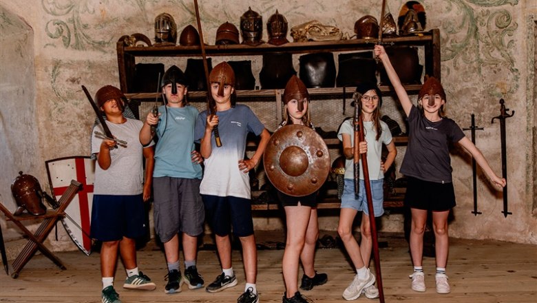 Group of children in medieval helmets and with weapons posing in a room with armor.