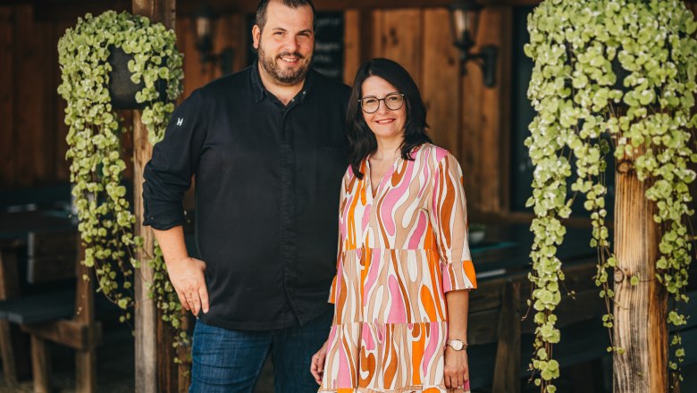 A man and a woman stand smiling next to each other in a rustic room with wooden walls and hanging plants.