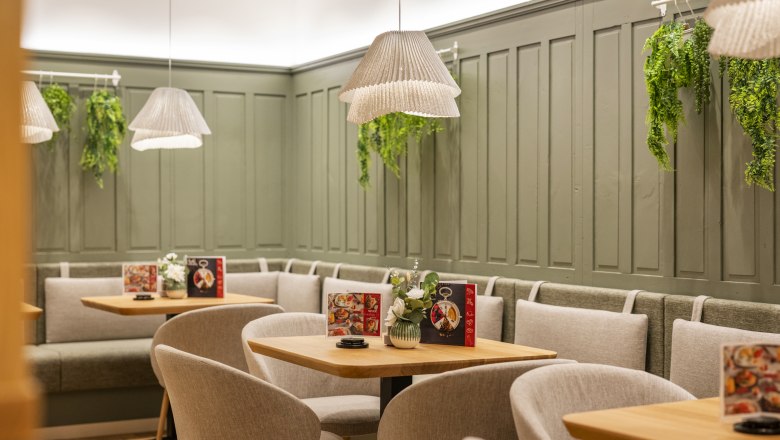 Cozy café area with wooden tables, grey chairs and green wall panelling. Hanging plants and lamps create an inviting atmosphere.