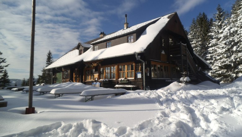 Snow-covered mountain hut with wooden benches in the foreground and fir trees in the background.
