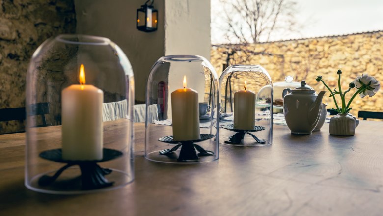 Three lighted candles under glass bells on a wooden table, next to a teapot and a vase with flowers.