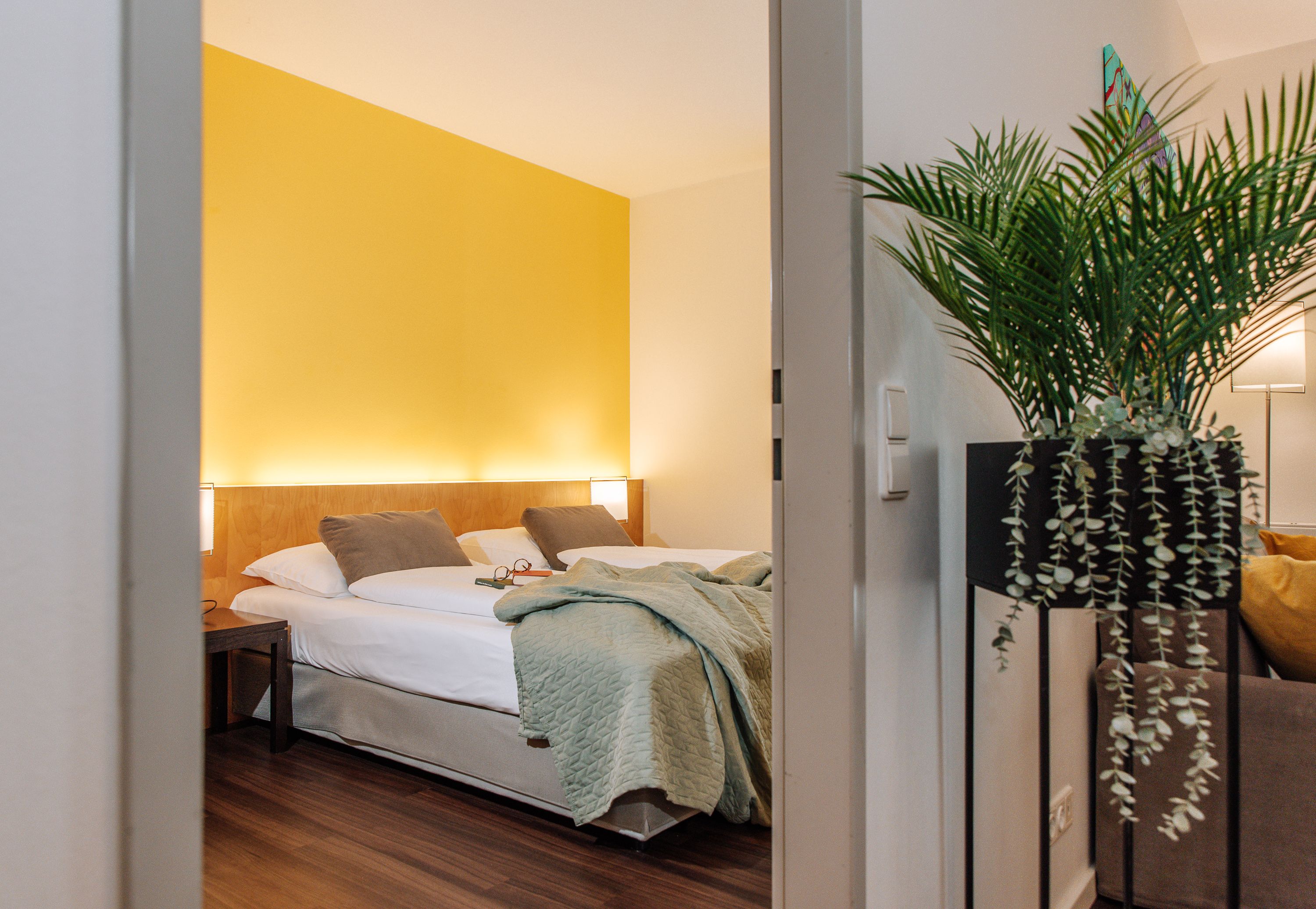 Hotel room with yellow wall, double bed and plant in the foreground.