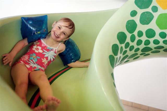 A laughing child with water wings slides down a green water slide.