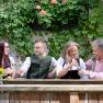 Four people in traditional dress clink glasses of wine outside in front of a wall covered in plants.