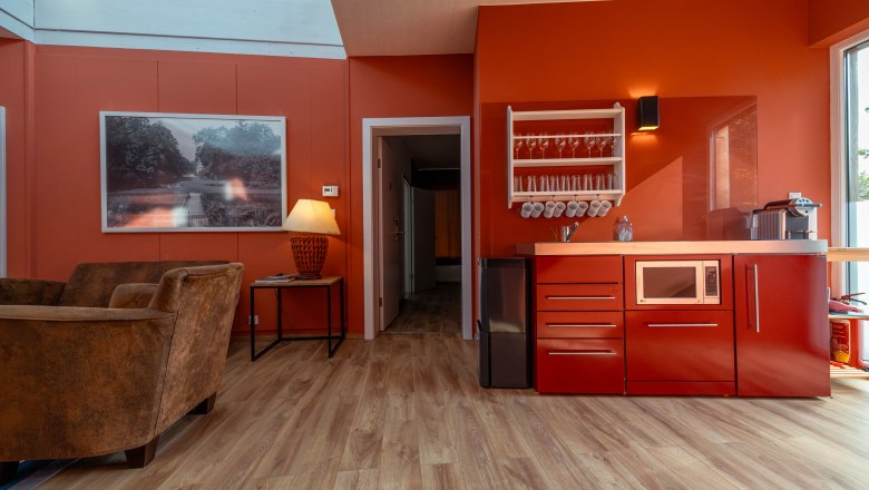 Interior view of a modern living room with red walls, a red kitchenette, brown armchairs and a large window.