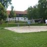 People playing volleyball on an outdoor sand court, surrounded by trees and a building in the background.