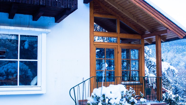 Winter view of the house with wooden veranda and snow-covered plants.