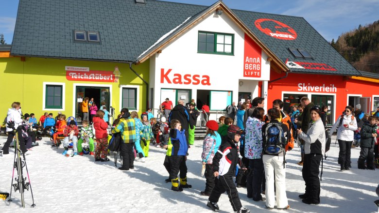 Crowd in front of a ski building with ticket office, ski rental and pond bar.