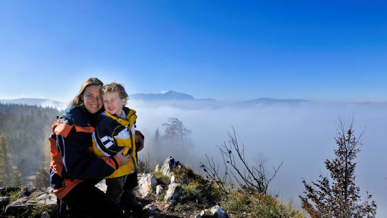 A woman and a child stand on a hill with a view of a sea of fog and mountains in the background.