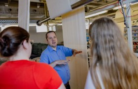 A man in a blue shirt explains something to two women on a factory floor.