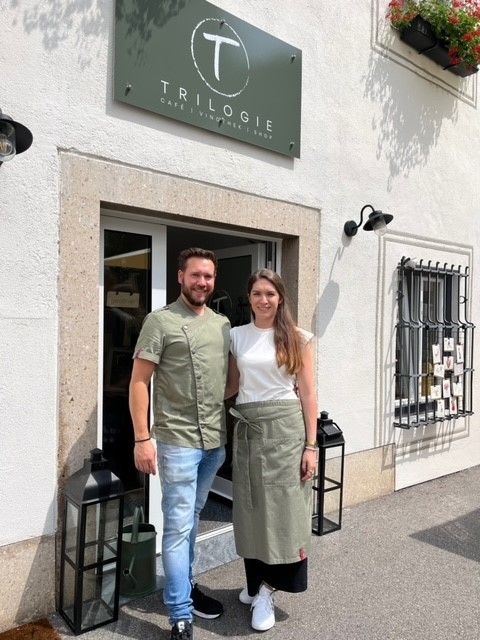 A man and a woman are standing outside a café called Trilogie.