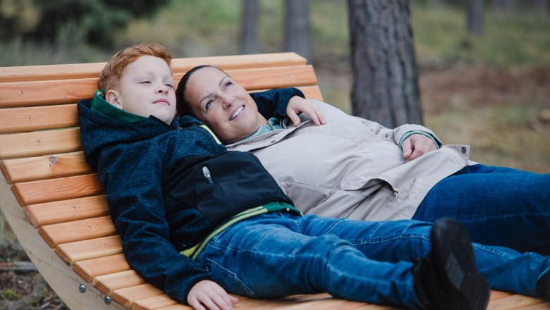 Two people lie relaxed on a wooden bench in the forest.